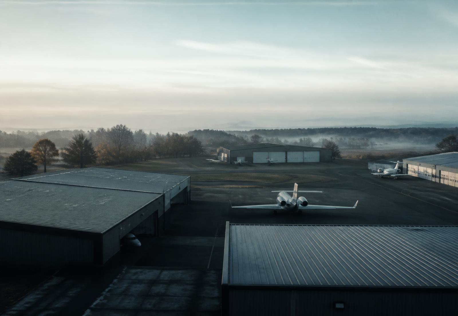 Private airfield at dawn with hangars and business aircraft on the apron in low mist